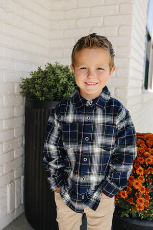 A young boy smiles while wearing a navy plaid button-up shirt made of soft cotton, standing in front of a brick wall and vibrant orange flowers.