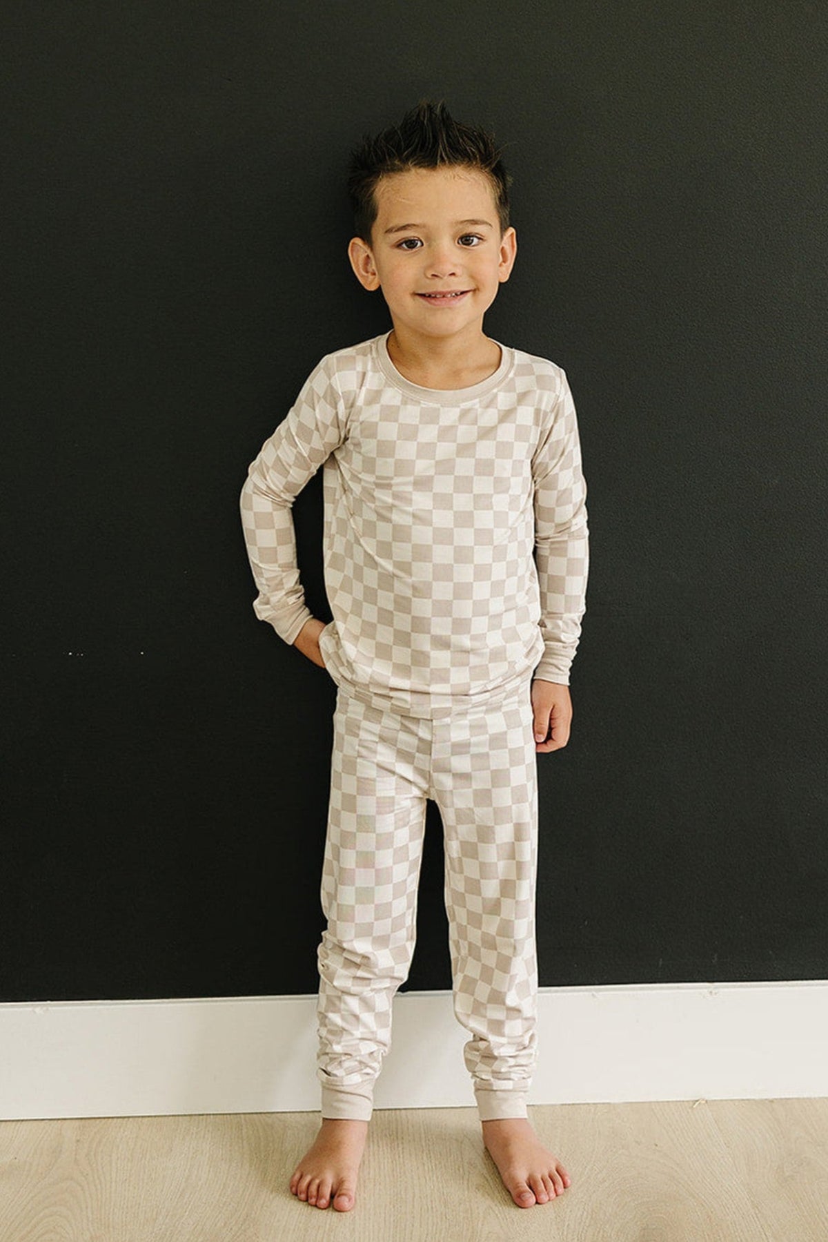 A child wearing a taupe checkered long-sleeve pajama set stands barefoot against a black wall.