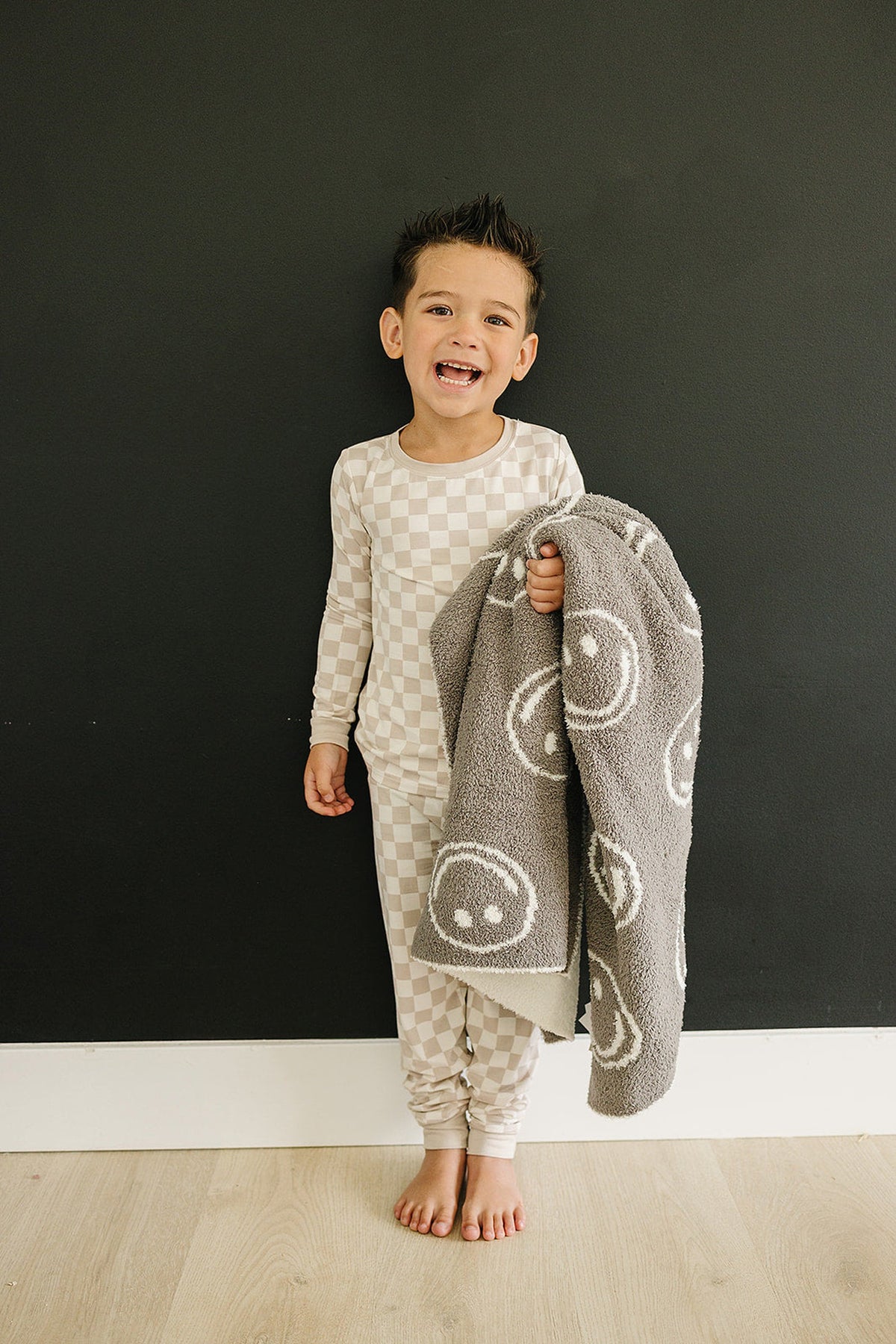 A child in a taupe checkered pajama set holds a gray blanket with smiley faces, standing against a dark wall.
