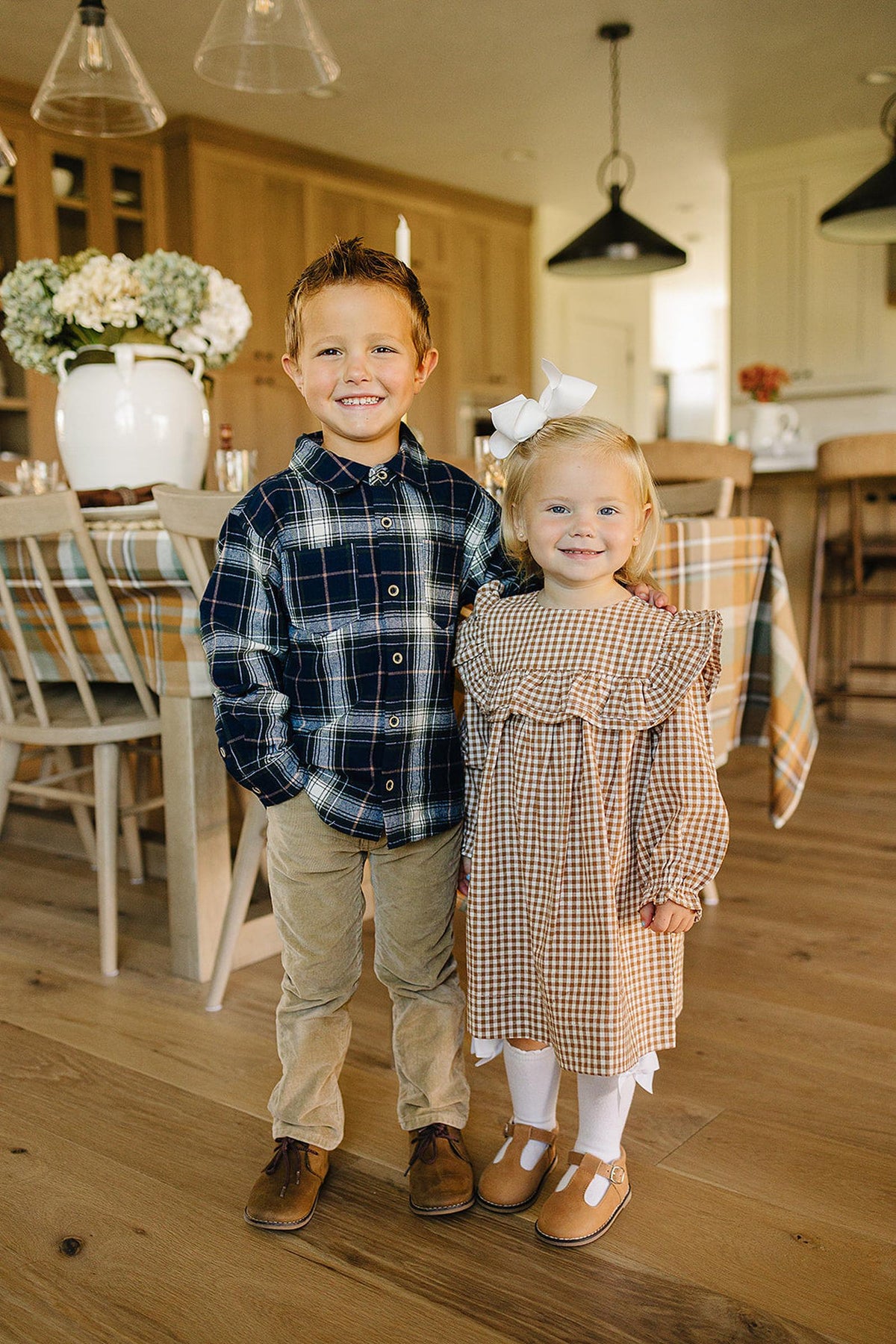 A smiling boy in a navy plaid button-up shirt stands beside a girl in a brown gingham dress with ruffles. They are in a cozy.
