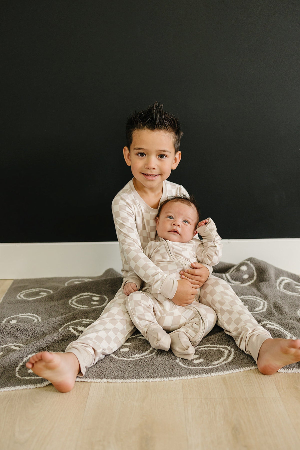 Two children in matching taupe checkered pajamas sit on a patterned gray rug against a dark wall.