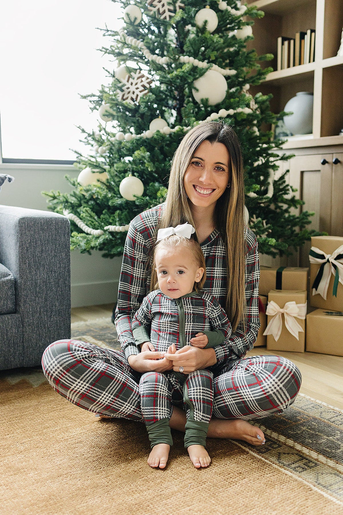 A woman and a toddler sit together on a rug, both wearing matching green plaid pajamas. The cozy setting features a decorated Christmas tree and festive gifts nearby.