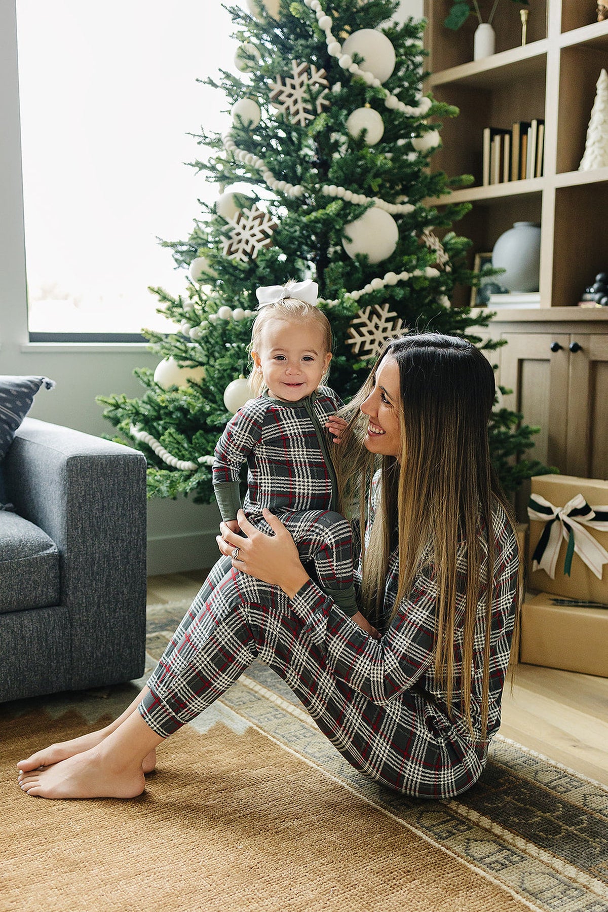 A mother and her child are seated together, both wearing matching green plaid bamboo zipper pajamas. The cozy living room features a decorated Christmas tree in the background.