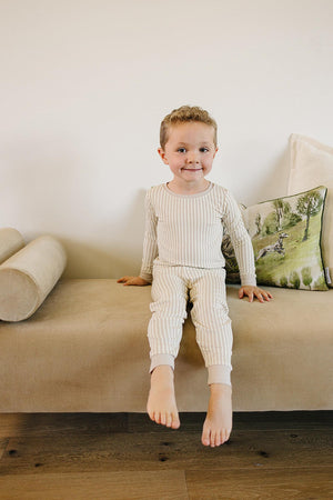 A smiling child sits on a beige couch, wearing a taupe striped pajama set made from soft, eco-friendly bamboo fabric.