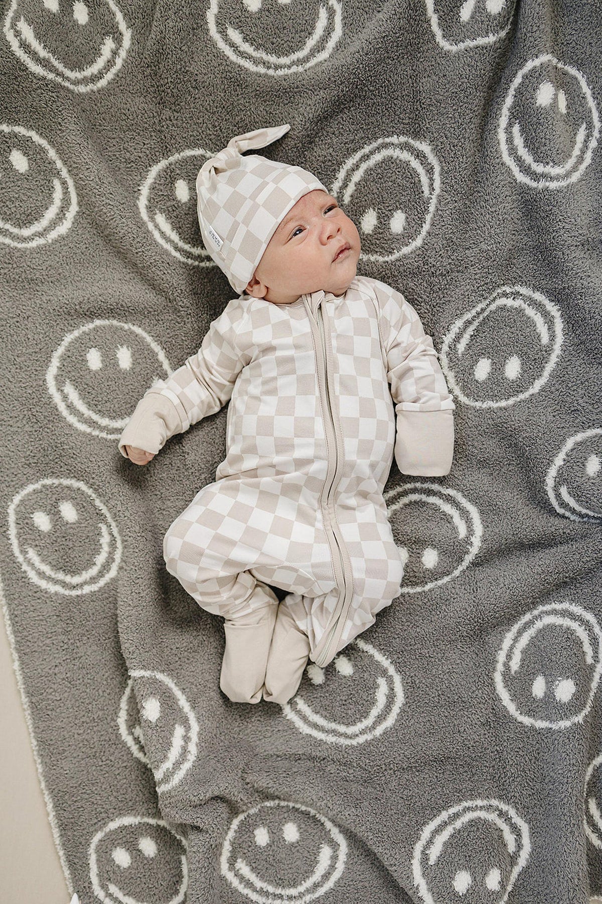 Baby in taupe checkered zipper pajamas and hat on a gray blanket with white smiley faces.