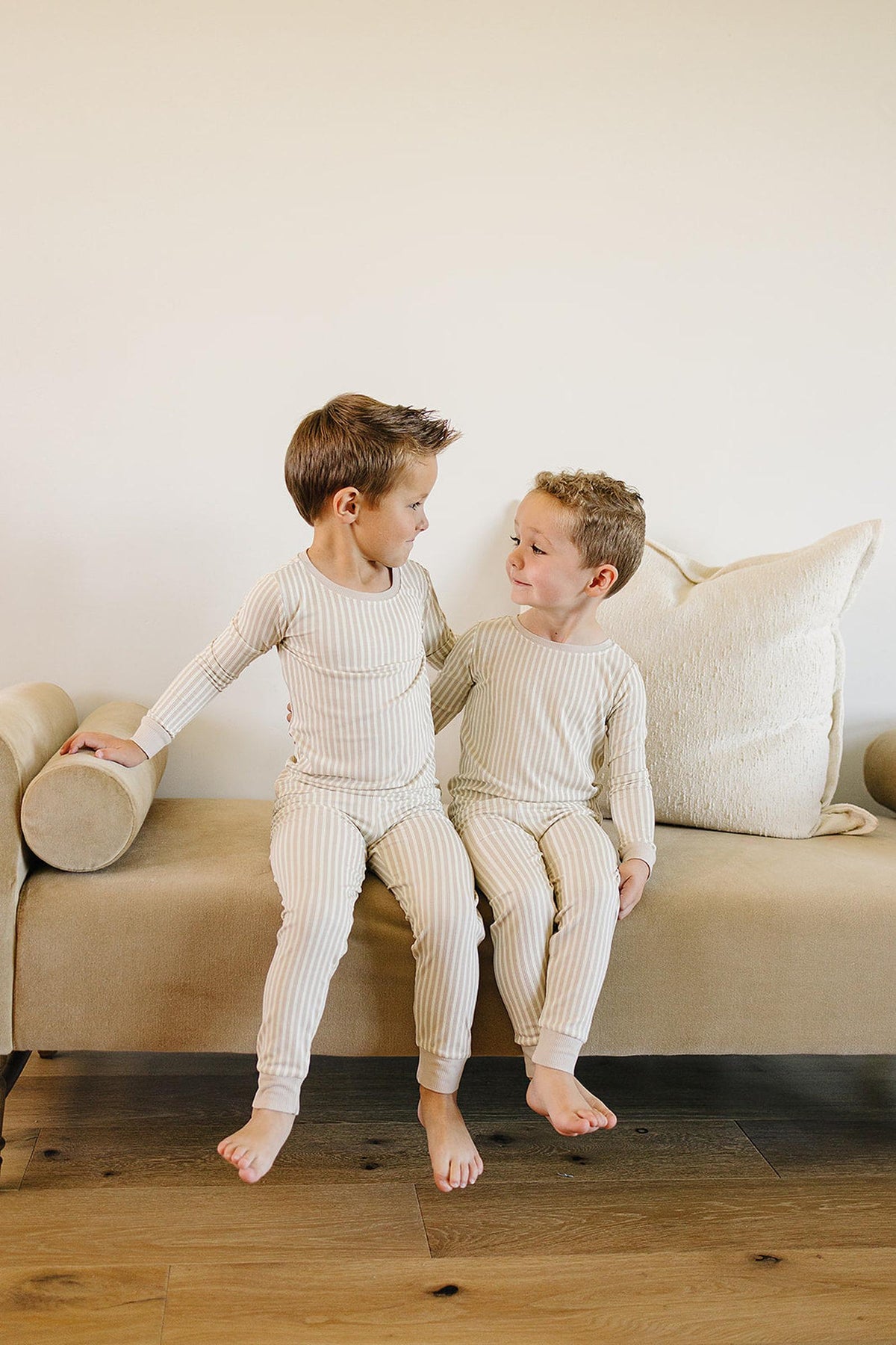 Two young boys sit on a beige couch, wearing cozy taupe striped bamboo pajamas, smiling at each other.