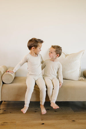 Two young boys sit on a beige couch, wearing cozy taupe striped bamboo pajamas, smiling at each other.