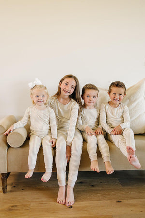 Four children sit on a beige couch, wearing matching taupe stripe ribbed pajamas made from soft bamboo fabric.