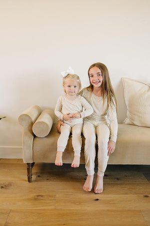 Two children sit on a beige couch, wearing matching taupe stripe ribbed pajama sets made from soft bamboo fabric.