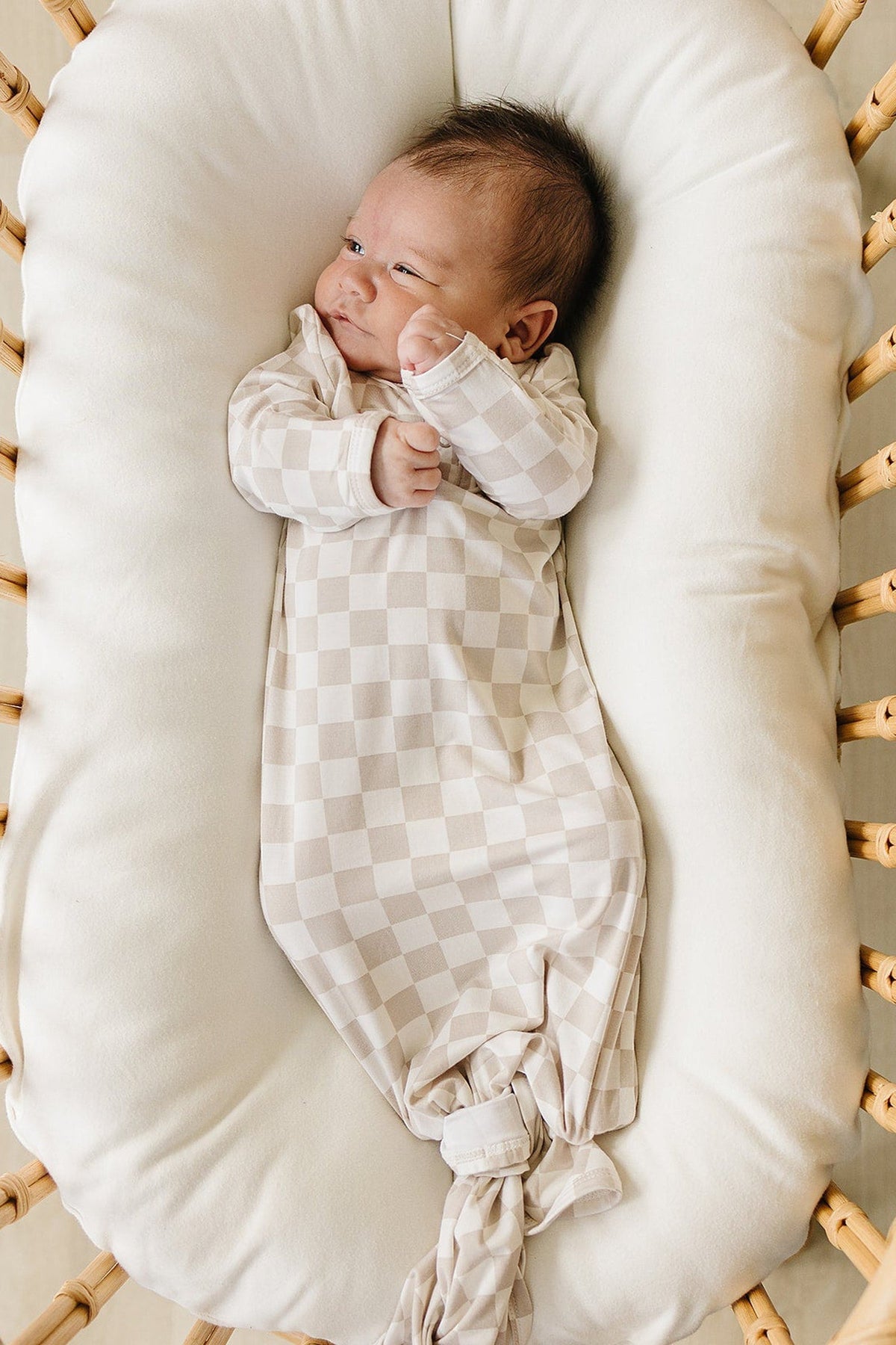 A cozy baby lies in a round bassinet, wearing a taupe checkered bamboo knot gown, with a soft white cushion.