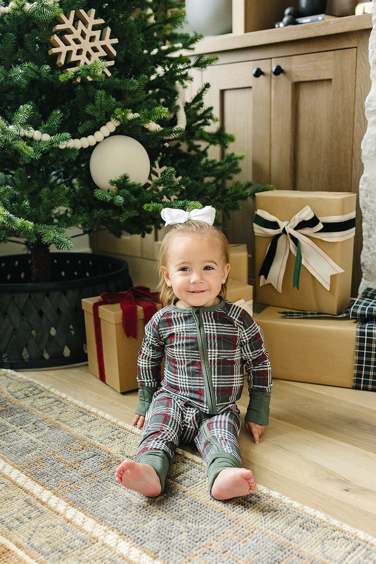 A toddler sits on a cozy rug near a decorated Christmas tree, wearing a green plaid bamboo zipper pajama. The setting includes wrapped gifts and a warm, festive atmosphere.