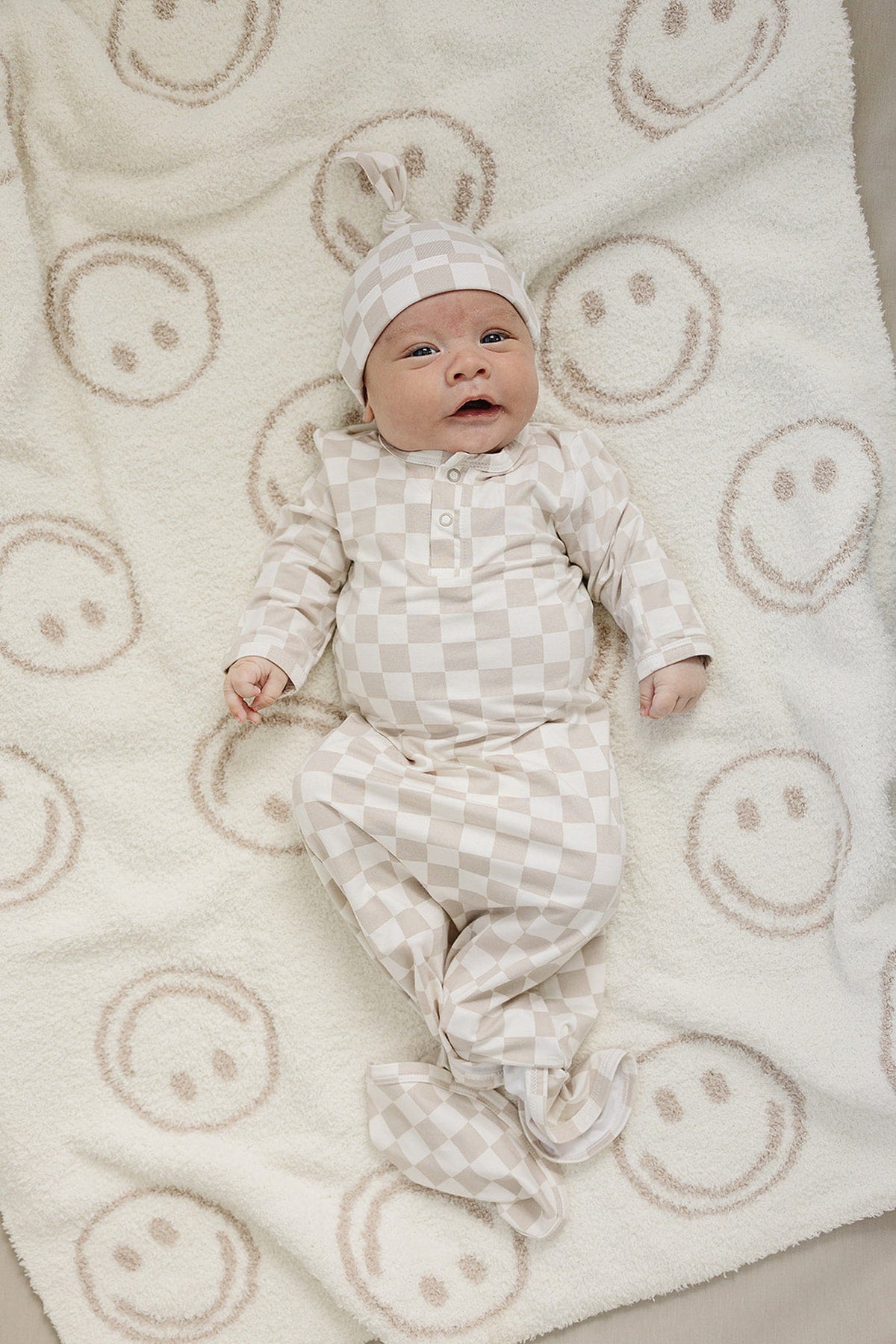 Newborn in a taupe checkered knot hat and outfit on a white blanket with smiley faces.