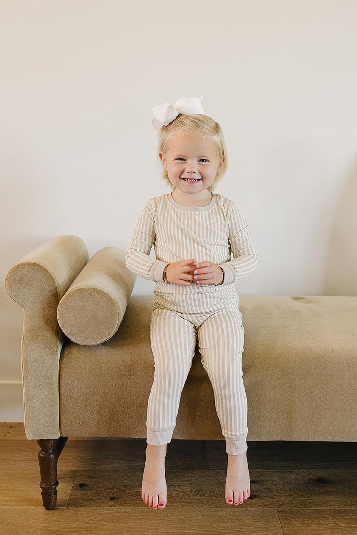 A smiling child sits on a beige couch, wearing a taupe stripe ribbed pajama set made of soft bamboo fabric. The outfit is perfect for cozy playtime or naptime.