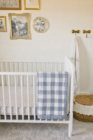 A dusty blue gingham muslin crib quilt drapes over the side of a white crib, set against a softly decorated nursery wall with framed artwork and a woven basket.