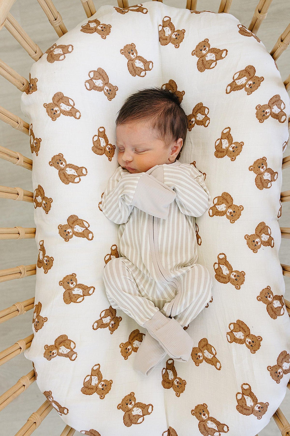 A sleeping baby in a taupe striped ribbed zipper pajama rests on a bear-patterned baby nest. The soft bamboo fabric ensures comfort and warmth for infants.