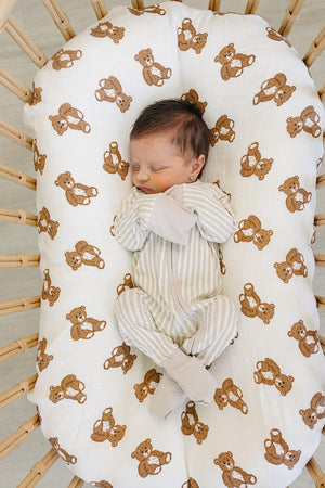 A sleeping baby in a taupe striped ribbed zipper pajama rests on a bear-patterned baby nest. The soft bamboo fabric ensures comfort and warmth for infants.