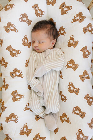 A sleeping baby in a taupe and cream striped ribbed zipper pajama rests on a bear-patterned muslin blanket. The pajamas are made from soft bamboo fabric, ensuring comfort and style.