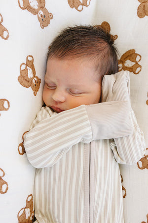 A peacefully sleeping newborn wears a taupe-striped ribbed zipper pajama, featuring soft bamboo fabric and long sleeves, resting on a cozy blanket adorned with teddy bear prints.