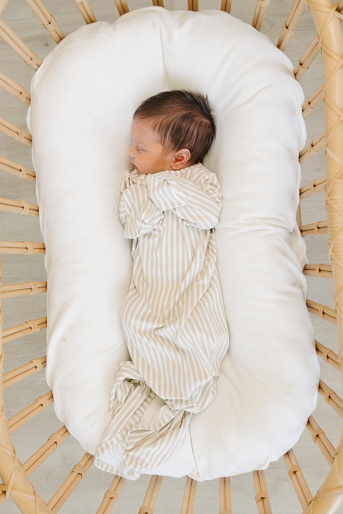 A newborn lies peacefully in a cozy bassinet, swaddled in a taupe striped ribbed gown made from soft bamboo fabric. The serene setting features a plush white cushion for comfort.