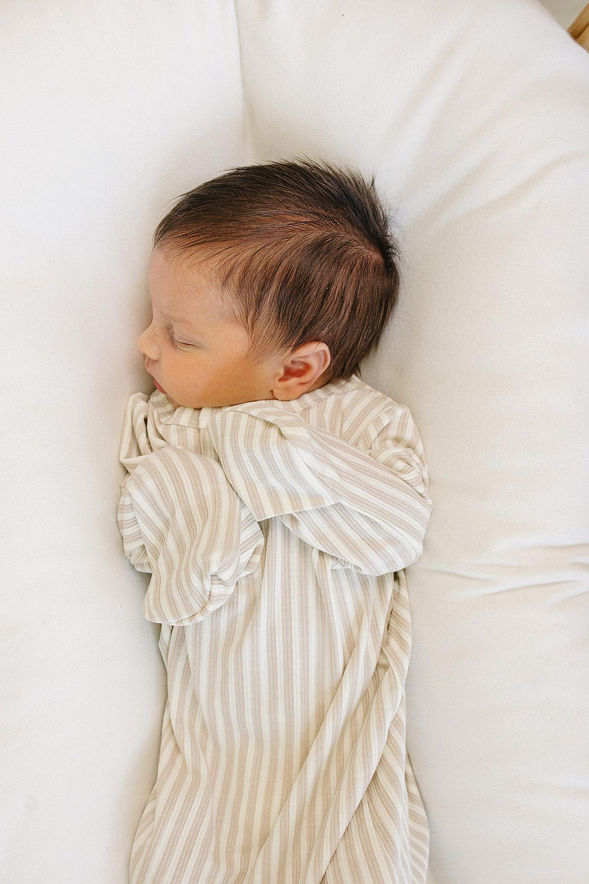 A peaceful newborn sleeps on a soft white surface, dressed in a taupe-striped ribbed gown. The gown features fold-over mittens, showcasing its comfortable and gentle bamboo fabric for delicate skin.