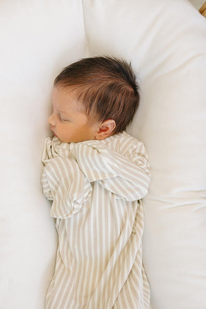 A peaceful newborn sleeps on a soft white surface, dressed in a taupe-striped ribbed gown. The gown features fold-over mittens, showcasing its comfortable and gentle bamboo fabric for delicate skin.