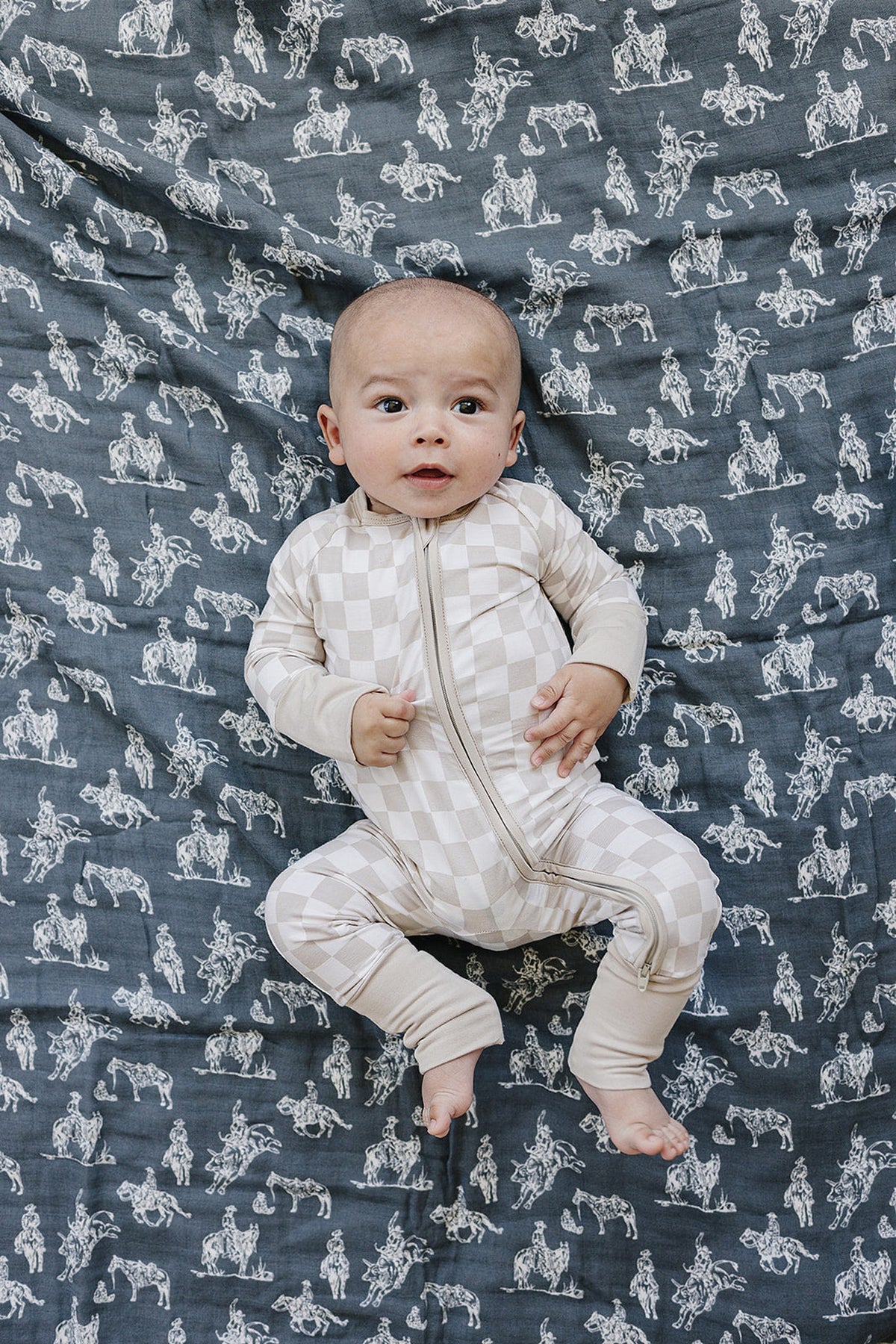 A baby in a beige checkered onesie lies on a gray quilt with white Wild West illustrations.