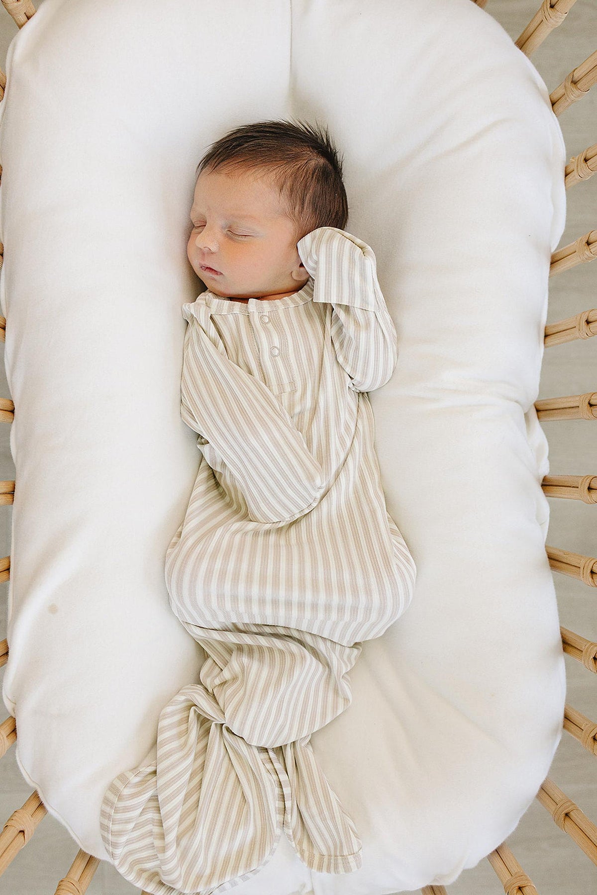 A newborn sleeps peacefully in a cozy, woven bassinet, dressed in a taupe striped ribbed knot gown made of soft bamboo fabric. The gown features folding mittens for added comfort.