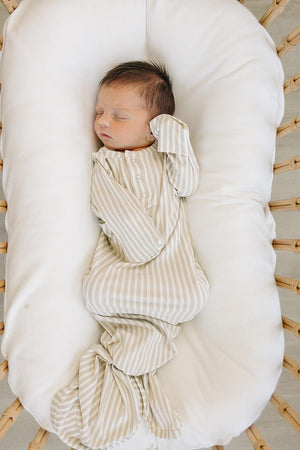 A newborn sleeps peacefully in a cozy, woven bassinet, dressed in a taupe striped ribbed knot gown made of soft bamboo fabric. The gown features folding mittens for added comfort.