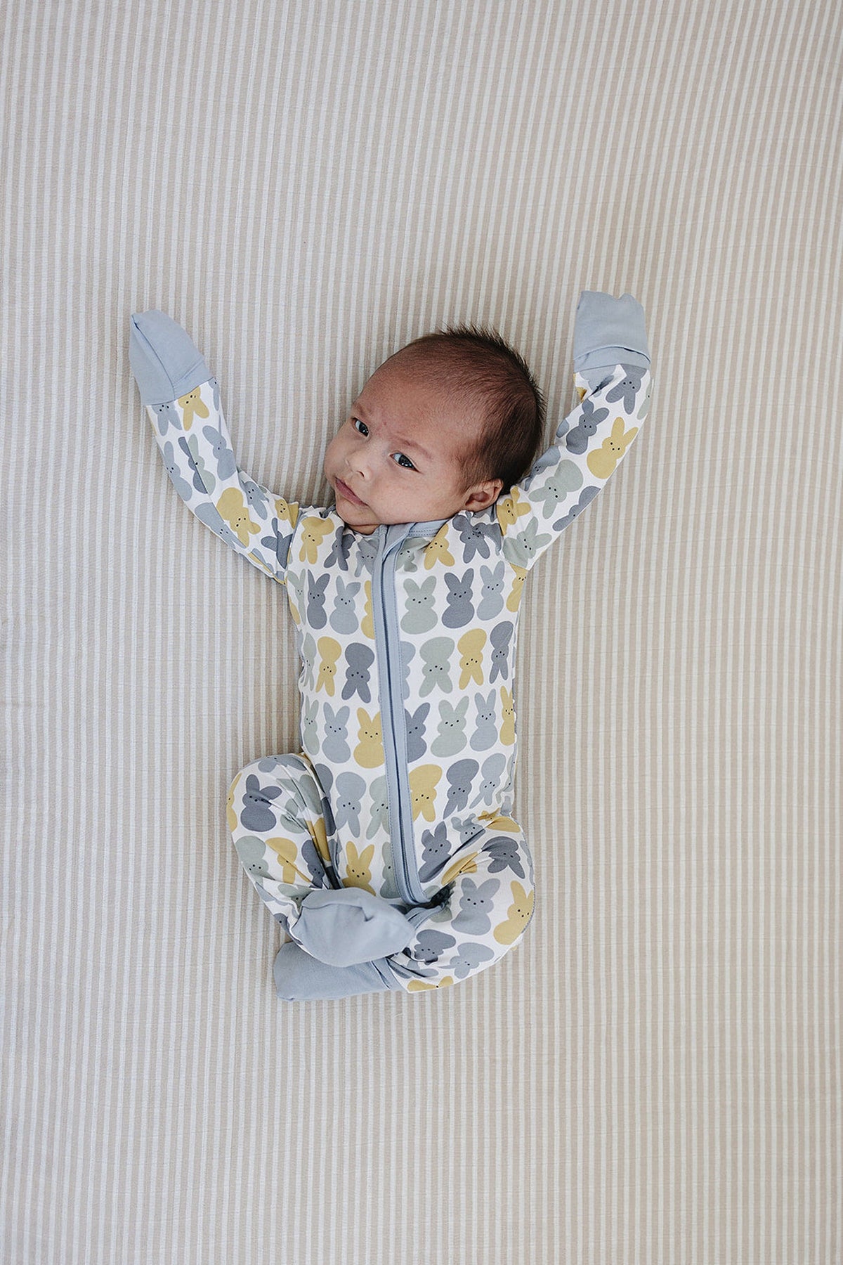 A baby wearing a patterned pajama with gray and yellow bunnies, lying on a striped beige surface.