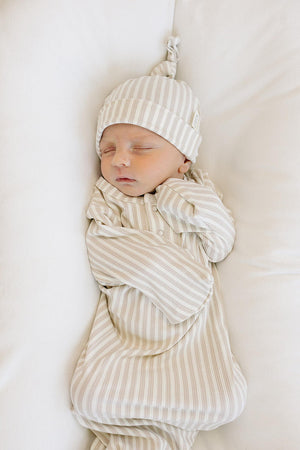 A sleeping newborn wears a taupe striped ribbed knot hat and matching outfit, resting on a soft white surface.
