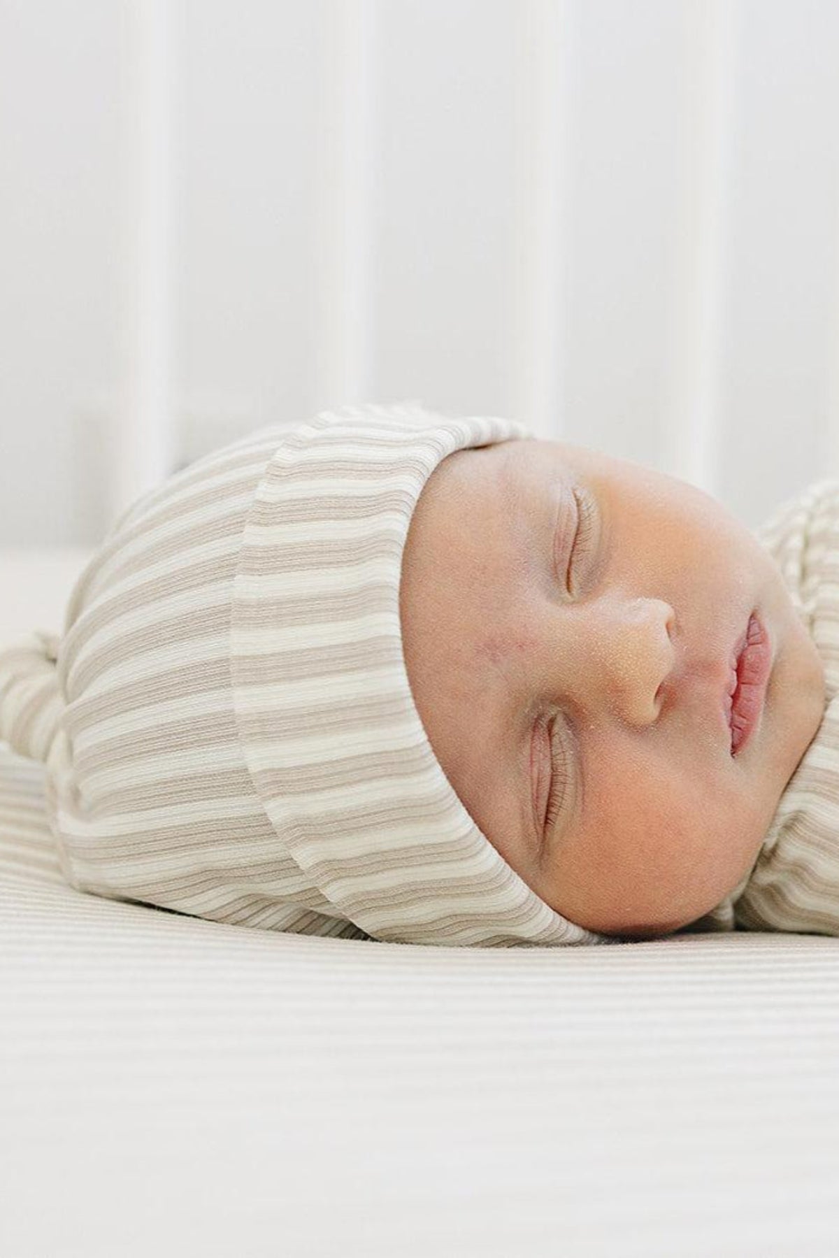 A sleeping newborn wears a soft taupe-striped ribbed knot hat, resting on a matching striped surface.