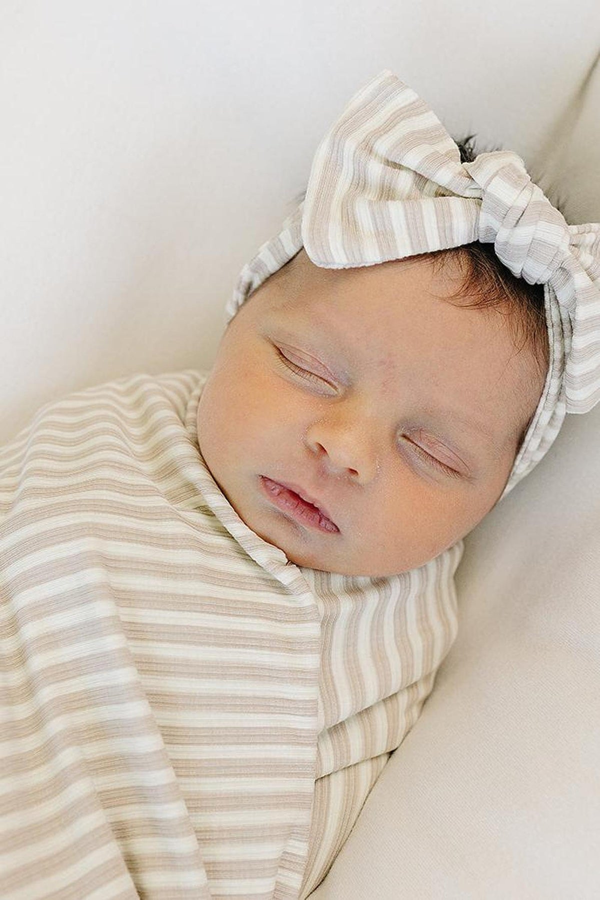 A newborn baby peacefully sleeps, swaddled in a soft, taupe-striped fabric. The baby wears a matching ribbed head wrap with a bow, showcasing the gentle, breathable bamboo material.