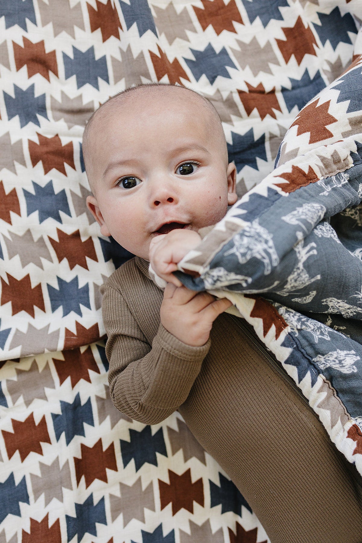 A baby in a brown outfit lies on a geometric-patterned quilt with blue, brown, and cream colors.