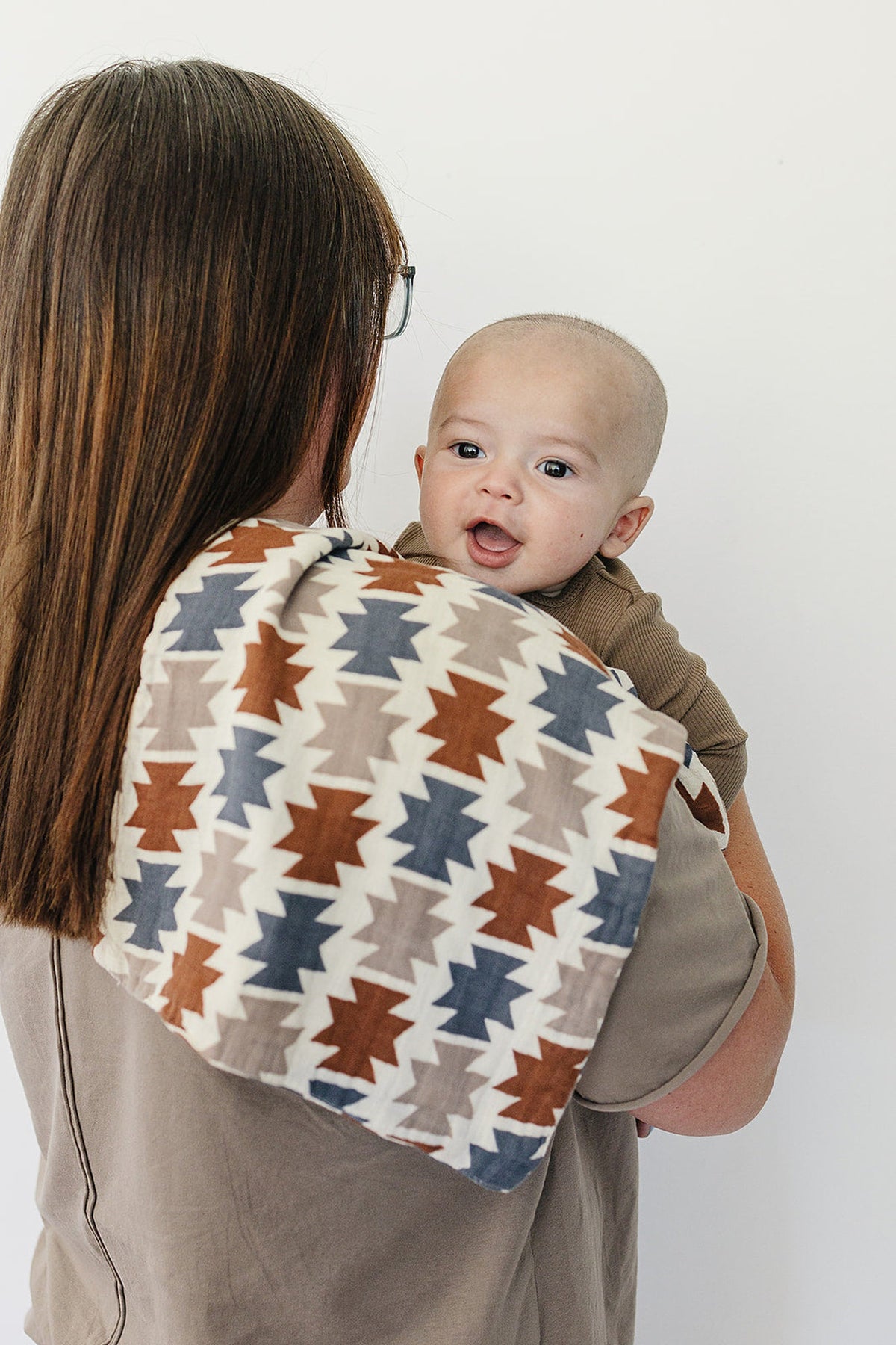 A parent holds a baby on their shoulder, showcasing a soft muslin burp cloth with a colorful Western Aztec pattern.