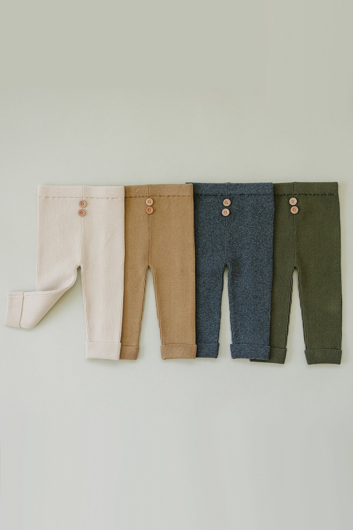 Four pairs of knit baby leggings in cream, brown, blue, and olive, each with two buttons at the waistband.