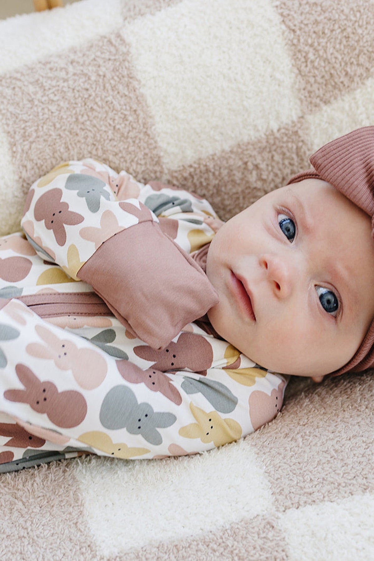 Baby in a multicolored bunny-patterned pajama with a dusty rose cuff, lying on a soft beige blanket.