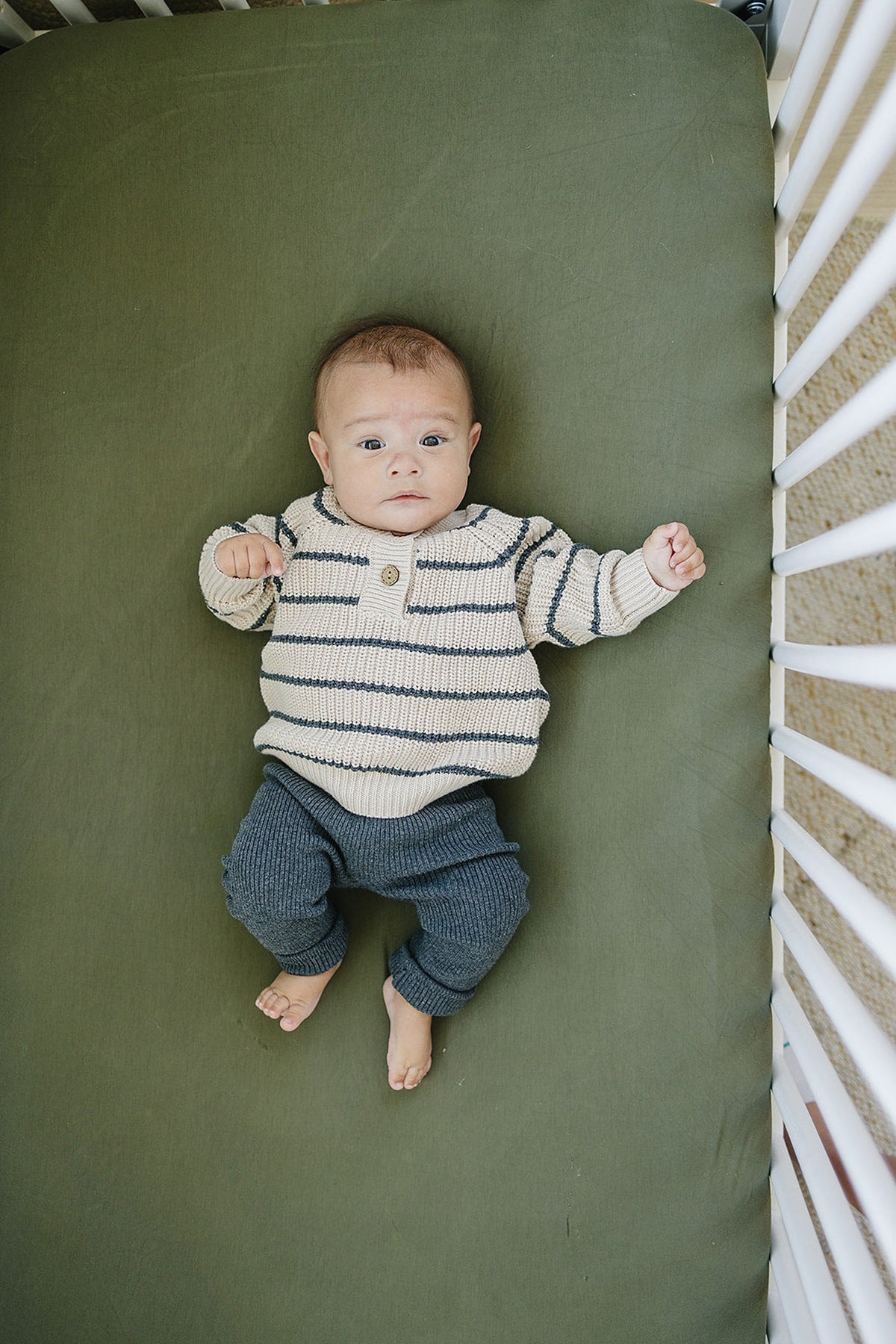 Baby wearing charcoal knit leggings and a striped sweater lies on a green crib sheet.