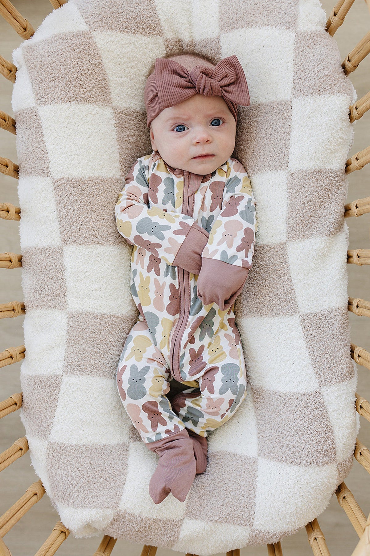 A baby in a bunny-patterned pajama and dusty rose headband lies on a soft checkered blanket.