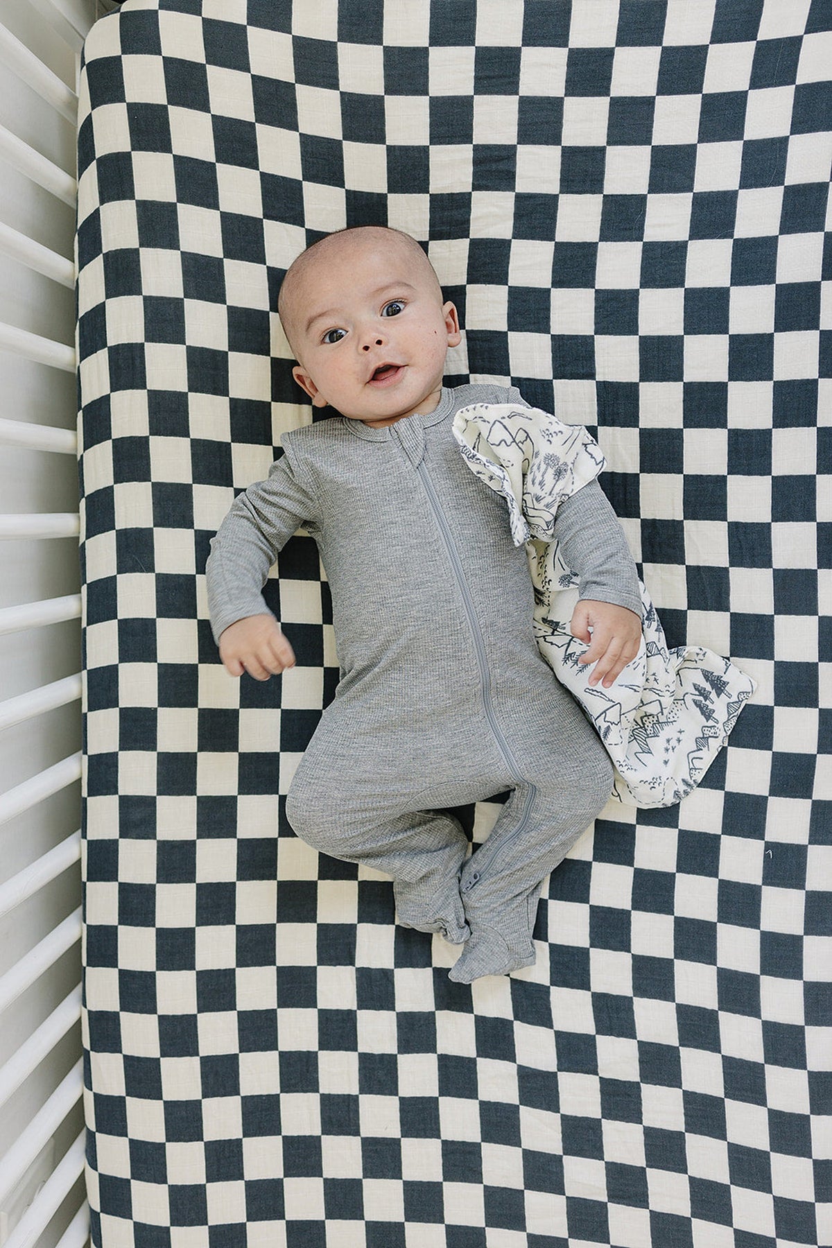 A baby in a gray onesie lies on a charcoal checkered muslin crib sheet, holding a patterned cloth.