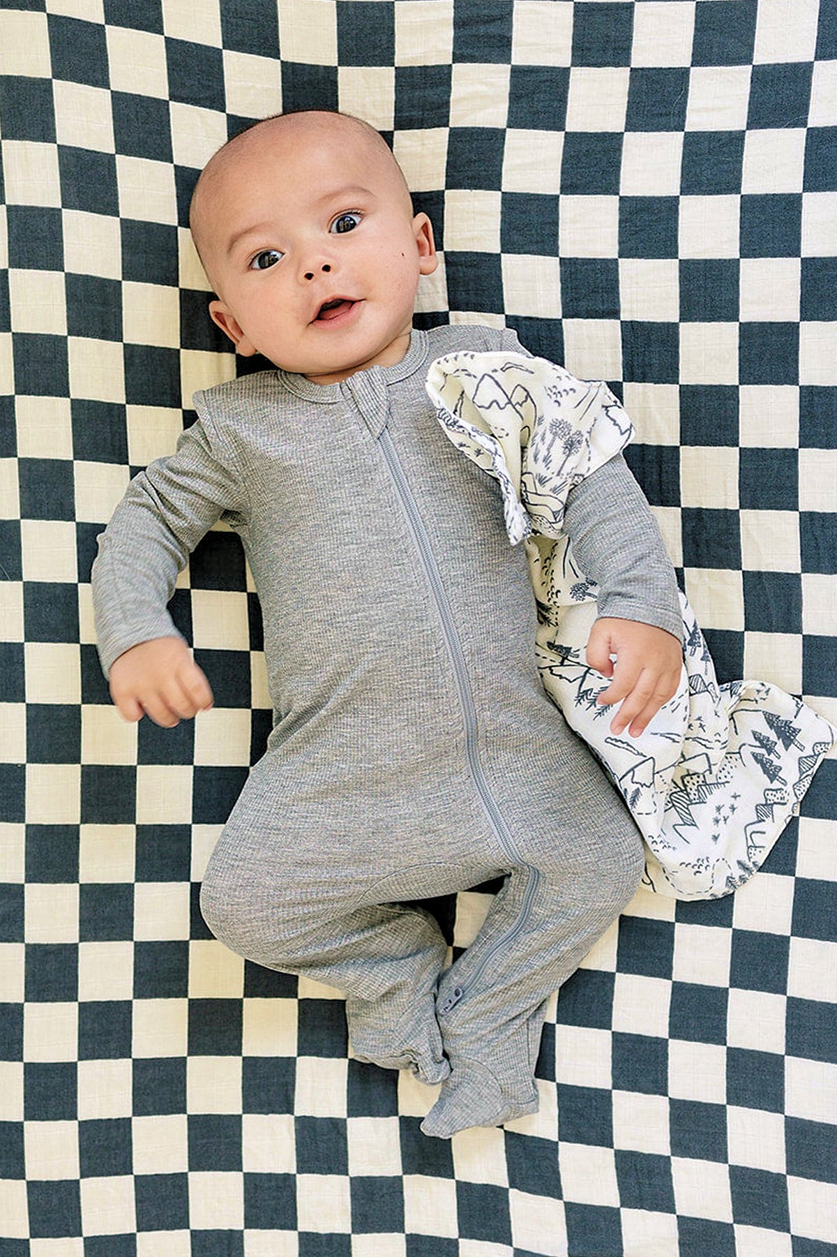 A baby in a grey ribbed zipper pajama lies on a checkered blanket, holding a patterned cloth.