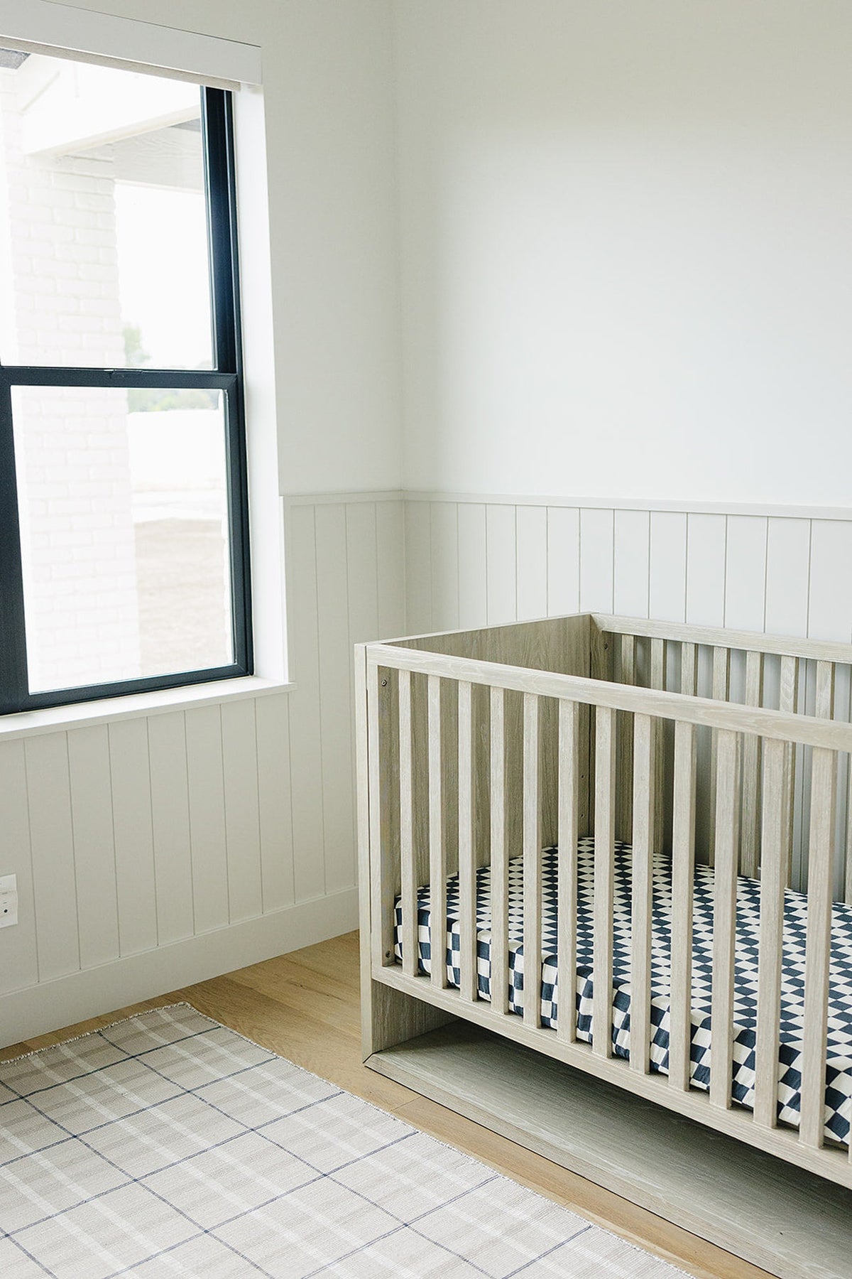 Wooden crib with a charcoal checkered muslin sheet in a bright nursery with white walls and a window.