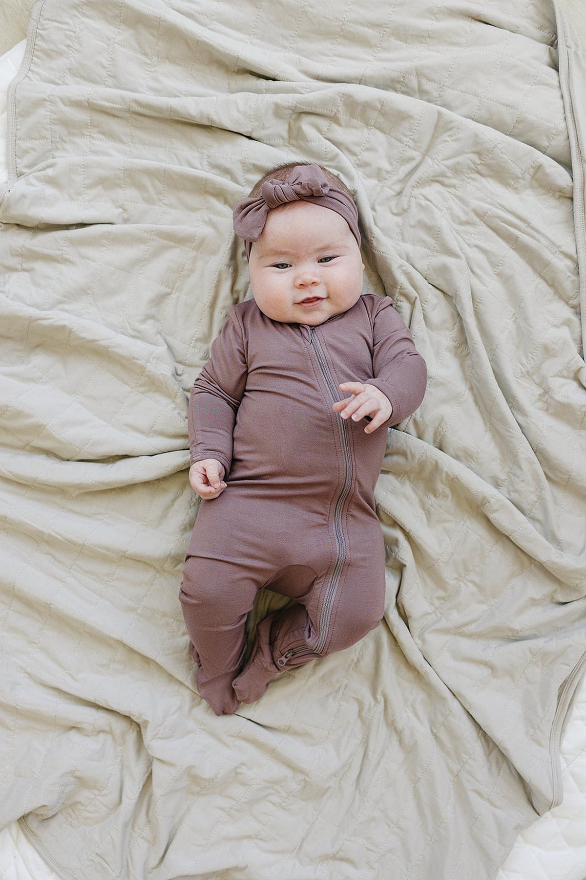 A baby wearing plum-colored footed pajamas with a headband lies on a soft cream quilted blanket.