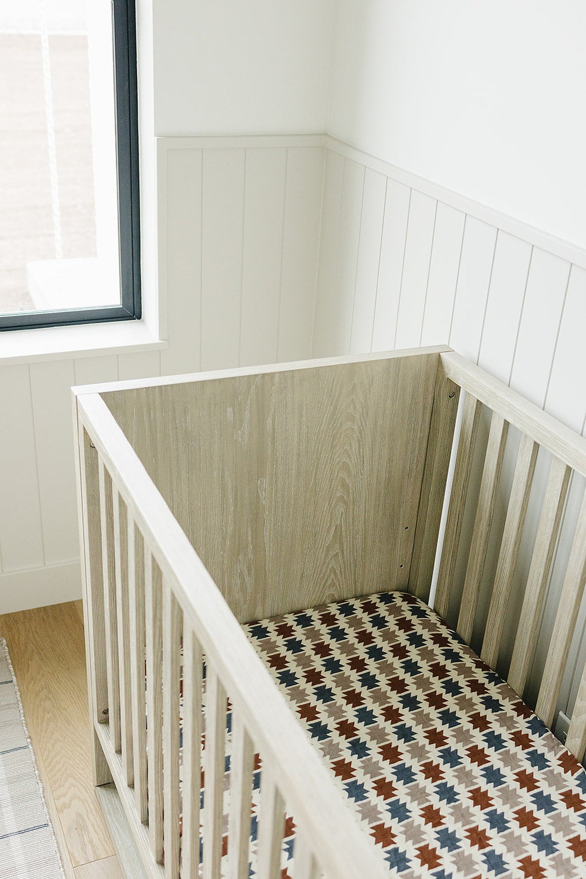 A wooden crib with a Western Aztec patterned muslin sheet in earthy tones, set against a bright, minimalistic room.