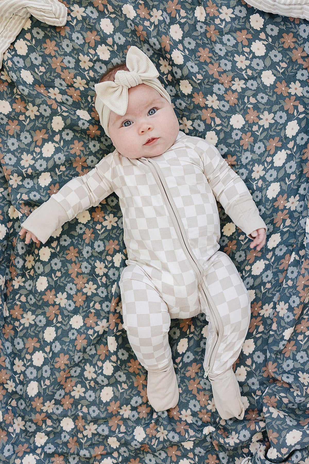 Baby wearing taupe checkered pajamas with a matching headband, lying on a floral-patterned blanket.
