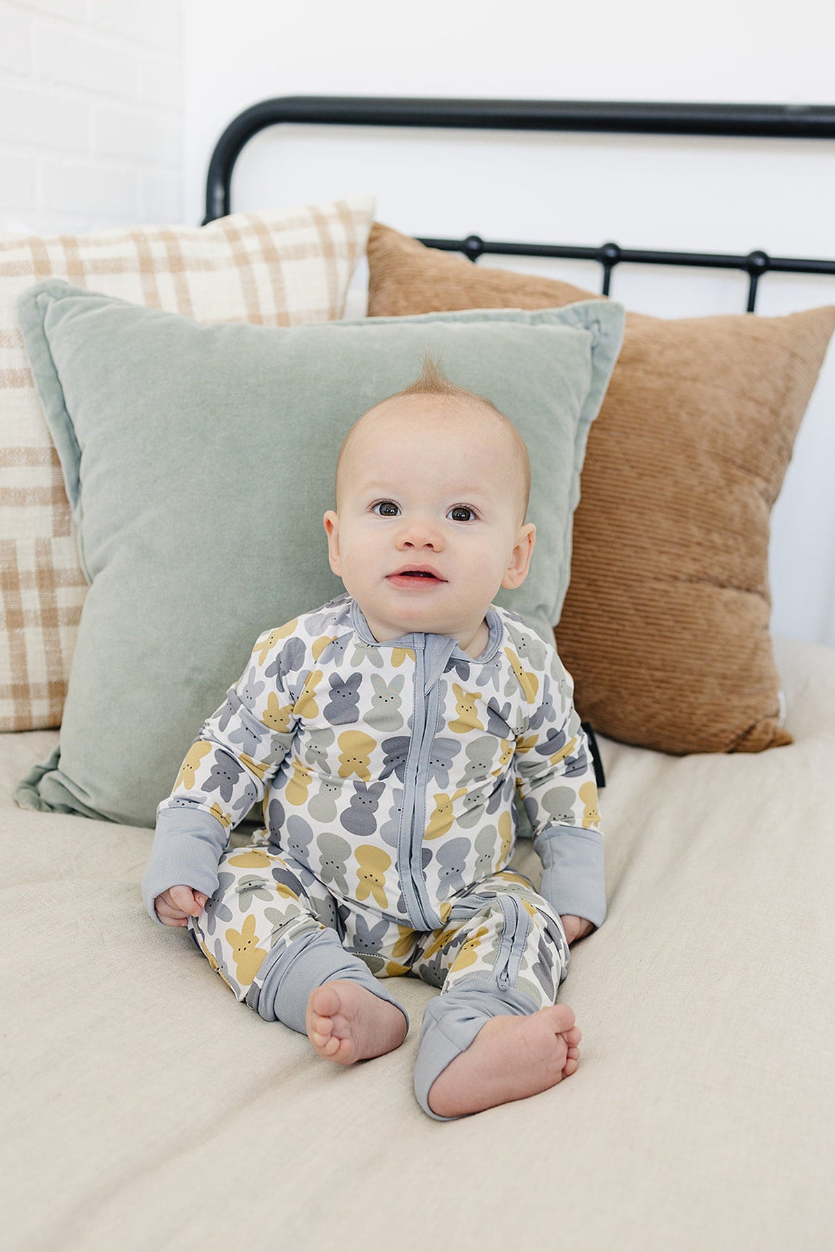 A baby wearing a gray and yellow bunny-patterned zipper pajama sits on a bed with pastel pillows.