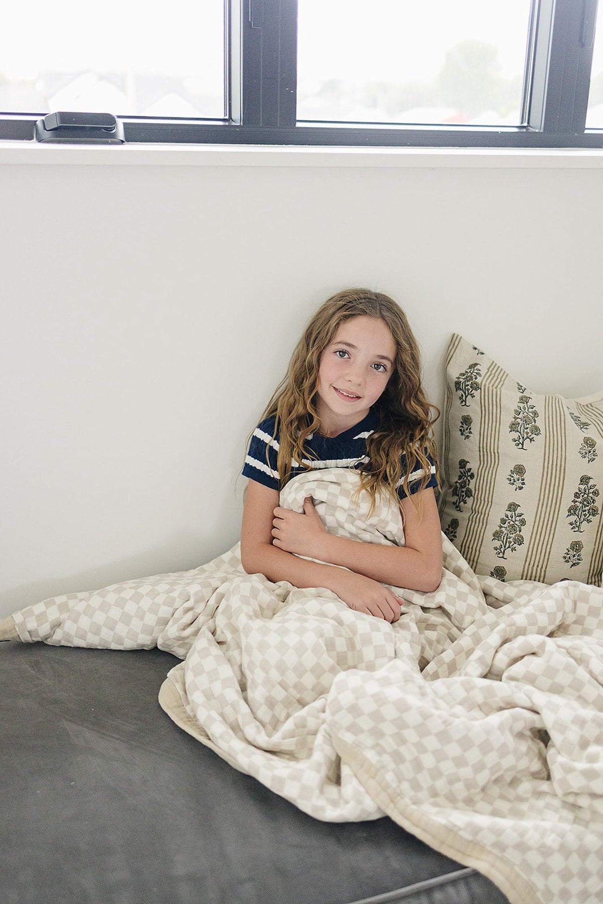 Young girl wrapped in a taupe checkered muslin quilt, sitting on a sofa with patterned cushions.