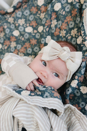 A sweet baby with large blue eyes wears a taupe checkered head wrap, surrounded by floral-patterned blankets in soft, muted.