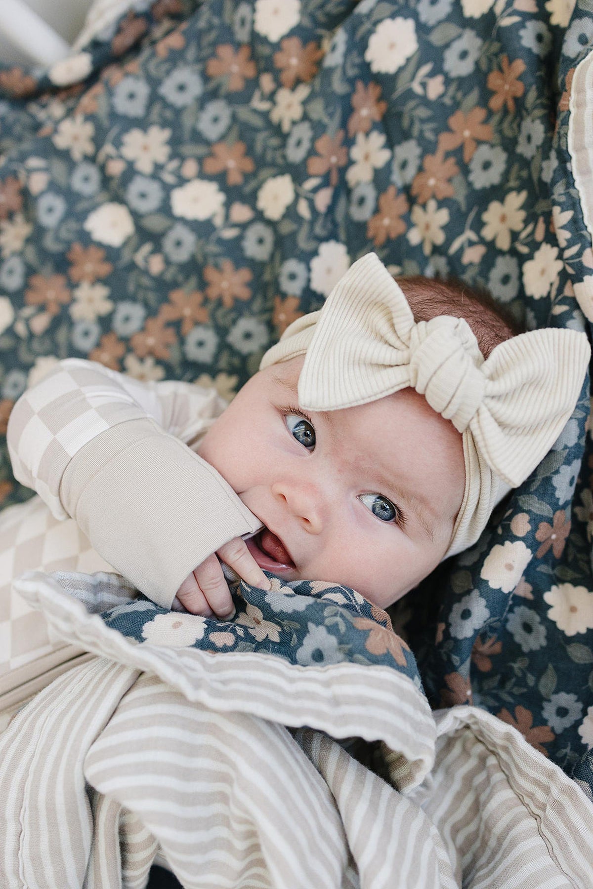 A baby wearing a taupe checkered pajama and cream bow headband lies on a floral blanket.