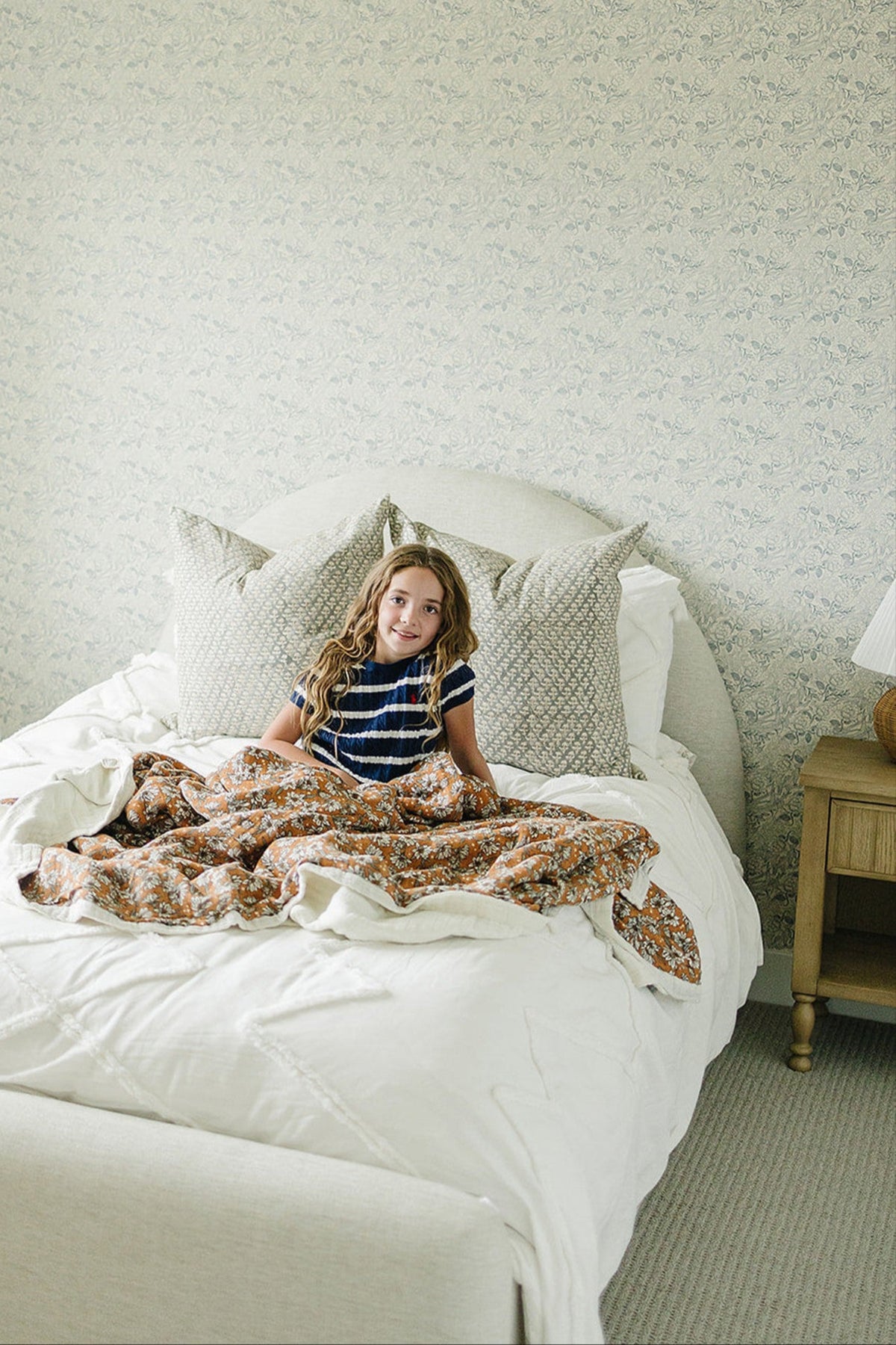 A young girl sits on a neatly made bed, wrapped in a floral muslin quilt with a light-colored comforter and.