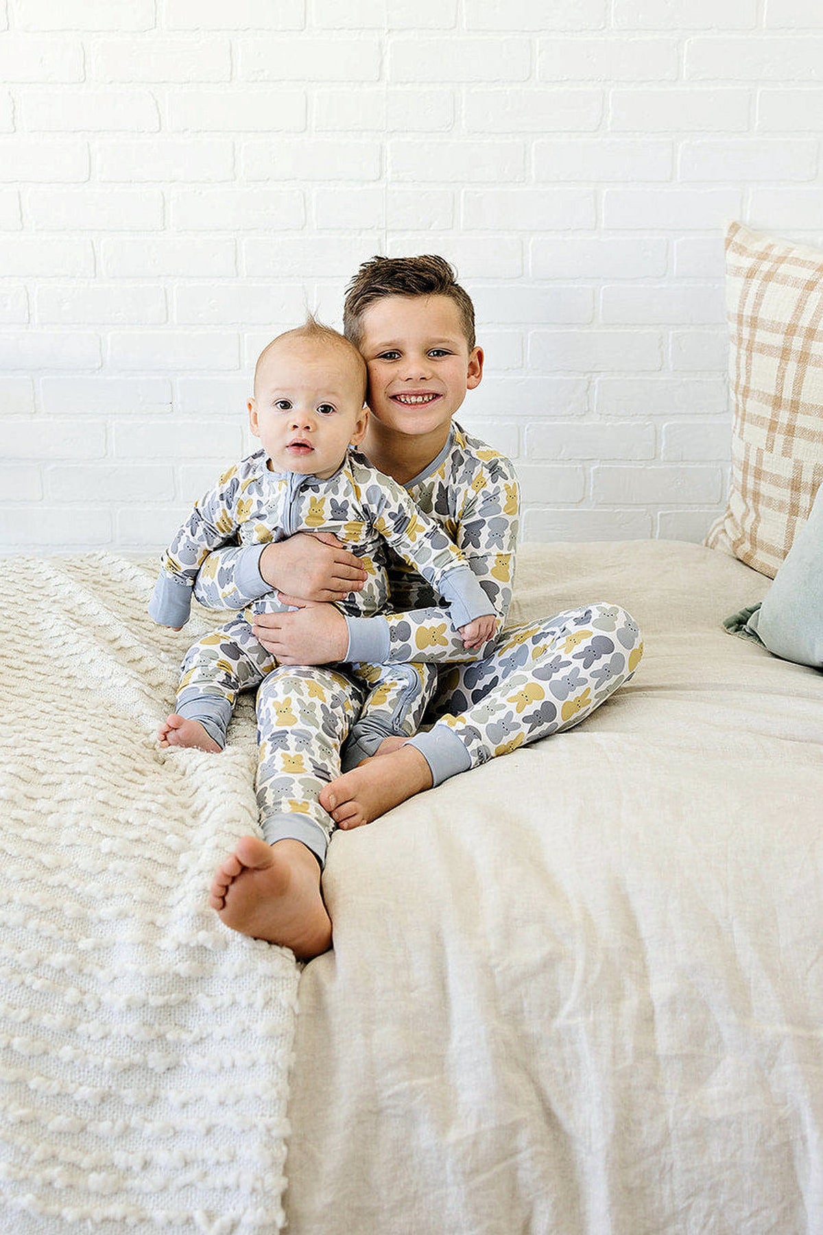 Two children wearing matching bunny-patterned pajamas sit on a bed against a white brick wall.